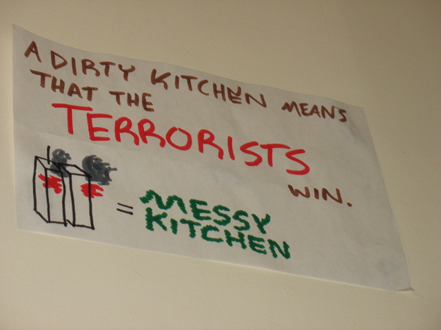 Sign posted very high in the kitchen: "A dirty kitchen means that the terrorists win. [Twin towers on fire] = Messy Kitchen"