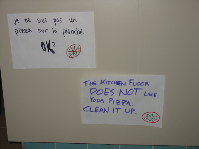 Two signs near the floor: "Je ne suis pas un pizza sur la planché. OK?" "The kitchen floor DOES NOT like your pizza. Clean it up."