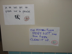 Two signs near the floor: "Je ne suis pas un pizza sur la planché. OK?" "The kitchen floor DOES NOT like your pizza. Clean it up."